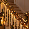 Warm white Christmas garland lights wrapped around a wooden staircase railing, decorated with green pine garlands and pinecones in a cozy indoor setting.