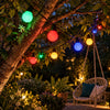 Multicolor LED globe string lights hanging in a garden tree, with a hanging wicker swing chair and plants in the softly blurred background.