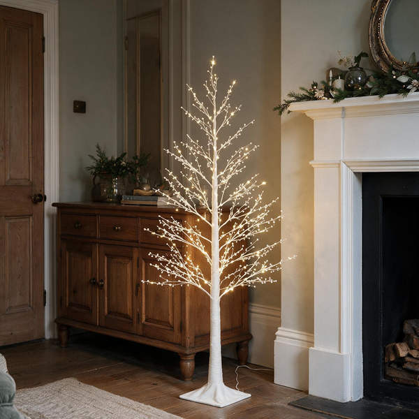 Pre-lit White Berry Twig Tree glowing softly beside a traditional fireplace and mantlepiece.
